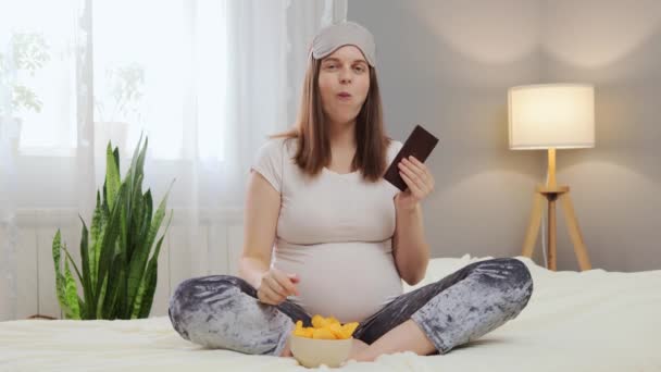 ブラウンは 妊娠中に美味しいジャンクフードを楽しんでチップとチョコレートのボウルがいっぱいの彼女のベッドに座っているパジャマの美しい若い白人妊婦をハイレッド — ストック動画