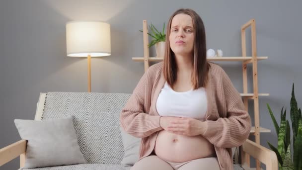 不健康なダークハイレッド妊娠中の女性は ソファーに座ってカジュアルな服を着て 彼女の腫瘍をストロッキングし 妊娠の最後の数ヶ月間は気分が悪くなっていると感じています — ストック動画