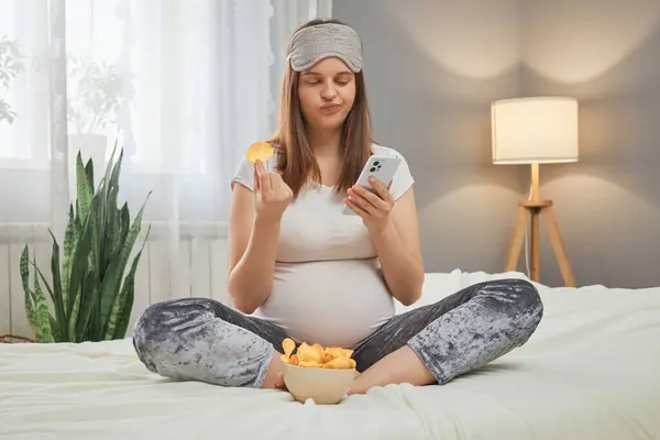 Puzzled Attractive Pregnant Woman Holding Smartphone Eating Chips Reading Harm — Stock Photo, Image