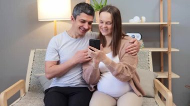 Çekici, neşeli, mutlu, hamile bir kadın ve yakışıklı bir adam oturma odasında birlikte oturmuş cep telefonu kullanıyor ve keyifli, ilginç içeriğin tadını çıkarıyorlar.
