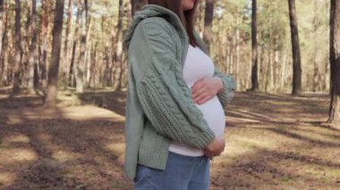 Tanımlanamayan hamile kadın parkta ya da ormanda dikilirken karnına dokunuyor yeşil örgü ören anne, ağaçta güzel doğanın tadını çıkarıyor.