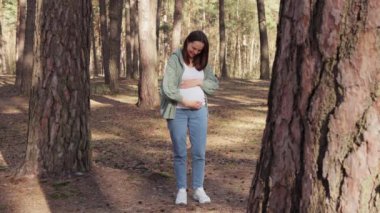 Gülümseyen neşeli hamile kadın, yeşil örgü kazak giyen, ormanda ya da parkta beklerken karnına dokunarak güzel doğanın tadını çıkarıyor.