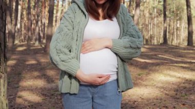 Kot pantolon giyen, yeşil örgü kazaklı, hamile bir kadın. Büyük göbeğini kucaklıyor, ormanda dinleniyor. Gelecekteki çocuğu temiz hava soluyor.