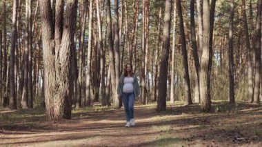 Son derece mutlu, neşeli, hamile bir kadın ormanda dinleniyor. Güzel doğanın ve çam ağacının tadını çıkarıyor. Parkta yürüyor, temiz hava alıyor ve günlük giysiler giyiyor.