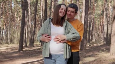 Sevimli, beyaz, hamile bir çift bahar ormanlarında rahatlarken mutlu, hamile bir anne ve kocası ormanda sevgi ve mutluluğu ifade ediyor.