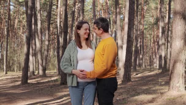 快乐美丽的年轻家庭孕妇和男人在森林里休息呼吸新鲜空气等待着父亲拥抱妻子的大肚子爱妻的夫妇一起享受怀孕和快乐时光 — 图库视频影像
