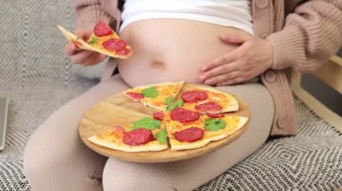 Tanımlanamayan hamile bir kadının bir dilim pizzanın tadını çıkarması çıplak karnını okşaması evde sağlıksız atıştırmalıklar yemesi.
