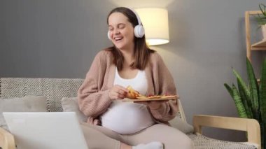Hamilelik yaşam tarzı. Mutlu hamile, beyaz, mutlu kadın dizüstü bilgisayar kullanıyor, internetten film izliyor, pizza yiyor, kanepede oturuyor ve gülüyor.