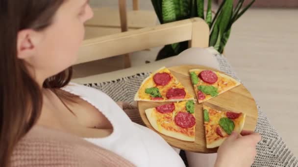 ブルネット妊娠中の少女は 居心地の良いリビングルームでソファーに座っている妊娠中にジャンクフードを楽しんで満足のいくビットスライスでピザを食べます — ストック動画