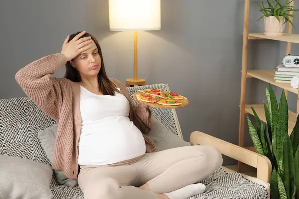 Dissatisfied pregnant woman sitting on couch with crossed legs touching her head feeling unwell overeating holding plate with pizza suffering after junk food dinner