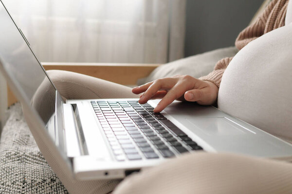 Relaxed unrecognizable unknown pregnant young woman using laptop at home sitting on couch typing on keyboard using computer for remote work during pregnancy