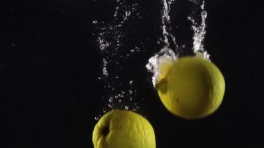Taze meyve sarısı limonlar suya dökülüyor. Siyah arka planda sıçrama var.