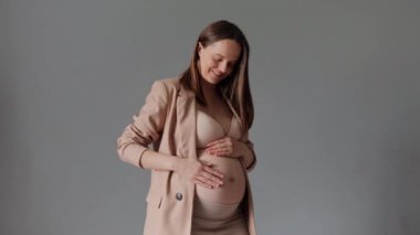 Kahverengi saçlı, bej ceketli, sırıtan hamile bir kadın göbeğini okşayarak gri arka planda tek başına duruyor.