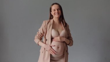 Bej iç çamaşırı ve ceket giyen neşeli, tapılası hamile kadın kameraya bakıp hamileliğinin tadını çıkarıyor.