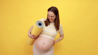 Çıplak karnıyla gülümseyen Kafkasyalı hamile kadın. Üstü açık yoga minderi giyiyor. Sarı arka planda izole edilmiş.