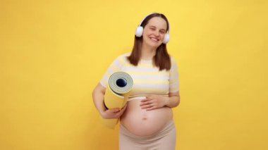 Çıplak göbekli, neşeli, beyaz bir kadın. Üstü başı açık, elinde karemat, sarı arka planda. Daha iyi spor çalışmaları için müzik dinliyor.