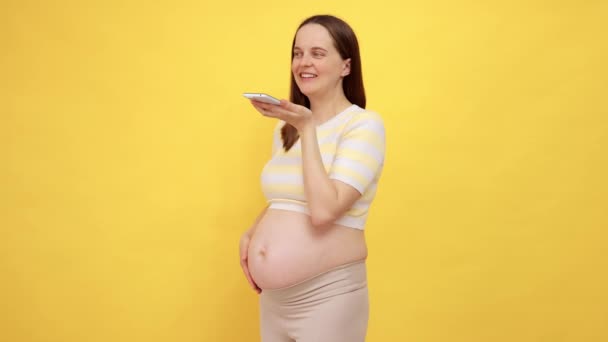 白种人孕妇裸着肚子 身穿休闲装 与黄色背景隔离 开心地微笑着 一边抚摸着她的肚子 一边记录着她给朋友的留言 — 图库视频影像