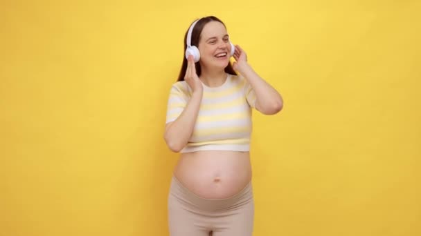 喜形于色的白种人孕妇 赤身露体 头戴黄色背景的休闲装 在耳机中欣赏着与未来婴儿的音乐 — 图库视频影像