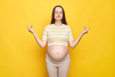 Calm relaxed Caucasian pregnant woman with bare belly wearing casual top isolated over yellow background practicing yoga trying to calm down standing with closed eyes clipart