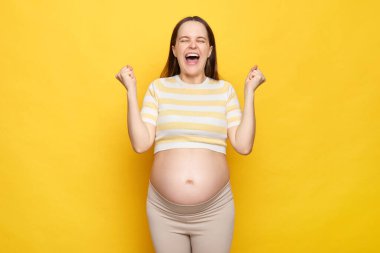 Aşırı derecede mutlu, aşırı mutlu, çıplak göbekli, sarı arka planda, üstü açık giyinmiş, beyaz bir kadın. Başarılı ultrason muayenesini kutluyor.