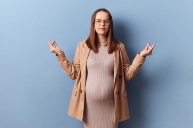 Calm relaxed pregnant elegant woman in beautiful dress and jacket posing isolated over blue background standing with mudra gesture meditating trying to calm down clipart