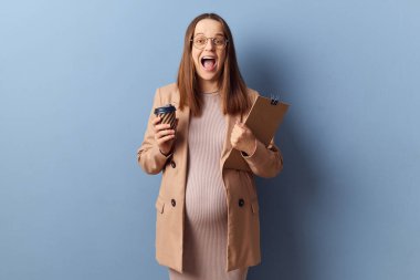 Aşırı mutlu, son derece mutlu, üzerinde elbise ve ceket olan hamile bir kadın elinde kahve, dosyasında mavi arka planda izole edilmiş heyecanlı bir yüz olduğunu iddia eden belgeler var.