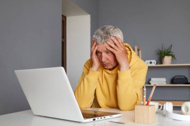 Evde dizüstü bilgisayar üzerinde çalışan üzgün yaşlı adam çok fazla çalışıyor şakaklarına masaj yapıyor bilgisayar yazılımlarıyla ilgili sorunları var mutsuz ve bitkin görünüyor.