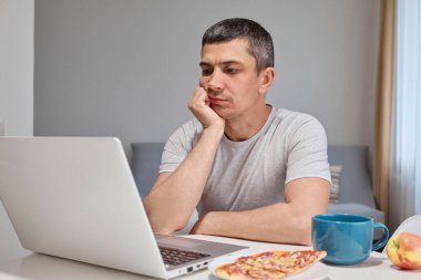 Sıkıcı, mutsuz, gri tişört giyen, evde dizüstü bilgisayarda çalışan, klavyede yazan arkadaşlarıyla sohbet eden, bilgisayarla uzaktan kumanda kullanan, mutsuz, beyaz bir adam.