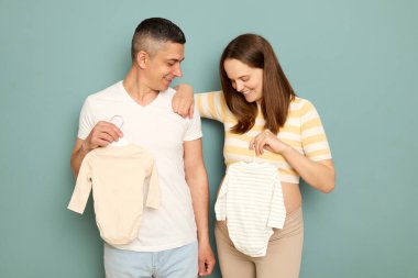 Hamile, beyaz, genç bir çift açık yeşil arka planda izole bir şekilde duruyor ve hamilelikleri boyunca bebek kıyafetleri seçiyorlar.