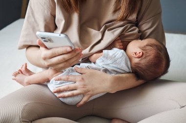Anonim kadın genç anne bebeğini ya da kızını emziriyor ve doğum izni sırasında internet üzerinden arkadaşlarıyla sohbet ediyor.