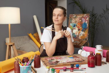 Sanat, yaratıcılık, hobi, iş ve yaratıcılık. Düşünceli kadın ressam atölyesinde oturup resim yapıyor ve yeni fikirleri düşünüyor. Hayal gibi bir ifadeyle başka yere bakıyor.