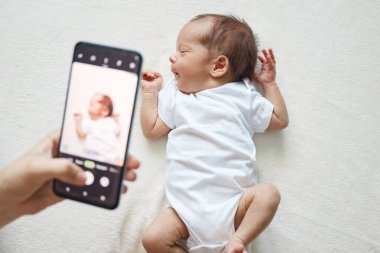 Anne, yeni doğmuş bebeğini akıllı telefonuyla beyaz çarşafta yatarak vuruyor. Bebeğinin cep telefonuyla fotoğrafını çekiyor. Oğlunun hayatının ilk günlerini çekiyor.