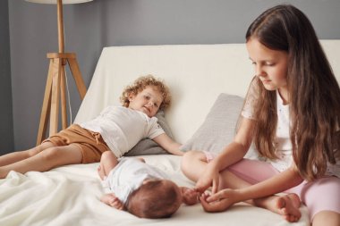 Büyük ablalar rahat, açık renk bir yatak odasında oturuyor. Küçük dalgalı saçlı kız kardeş kanepede uzanıyor. Kahverengi saçlı kız ise yeni doğmuş bebeği oynuyor.