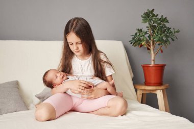 Evdeki kanepede oturan kahverengi saçlı kız ablamın elinde yeni doğmuş erkek kardeşiyle evde bebekle tanışmasını seyrediyor.