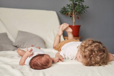 Dalgalı saçlı abla yeni doğan erkek kardeşiyle oynuyor. Kardeşiyle evde buluşuyor. Küçük bir çocukla beyaz kanepeye uzanıyor ve parmağını tutuyor.