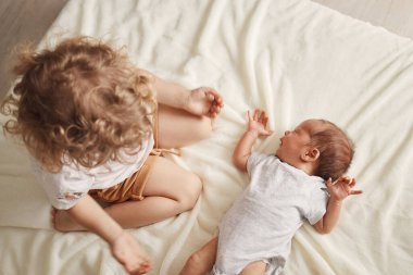 Top view of wavy haired little girl enjoying time spending with her newborn brother child looking at infant baby while playing with him on white soft sheet clipart