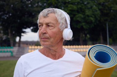 Güçlü ve sağlıklı kalmak için stadyum antrenmanlarına katılan yaşlı atletik adam kablosuz kulaklıkla yoga minderiyle dışarıda meditasyona hazırlanıyor.