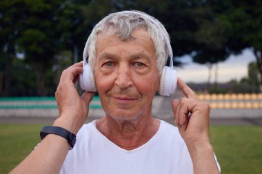 Şehir stadyumunda kulaklıklarla dikilmiş, kablosuz cihazına dokunan gülümsemesiyle kameraya bakan ve müzik dinleyen olgun bir beyaz adam.