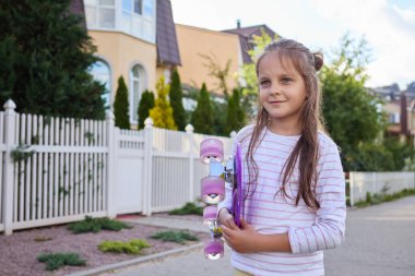 Kahverengi saçlı, günlük giysiler giyen, sörf tahtasını tutan, rüzgarlı bir günde şehir sokağında poz veren, aktif çocuklarda neşe bulan neşeli kadın çocuk.