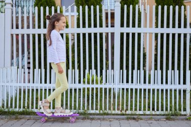 Kahverengi saçlı, aktif, günlük kıyafetlerle gezen, uzun panoya binmiş şehir sokaklarına bakan kaykaylı eğlenceli zamanların tadını çıkaran küçük bir kızın uzun kenar manzarası.