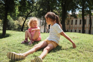 Sevimli, mutlu küçük kız kardeşler parkta yeşil tepede çimlerin üzerinde oturmuş neşeyle gülüyorlar yaz tatilini dışarıda geçirirken.