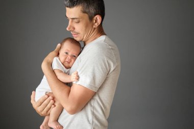 Sevimli beyaz bir baba, yeni doğmuş bebeğini kollarında tutuyor. Gri arka planda, beyaz tişört giyen genç adam reklam için fotokopi odası giyiyor.