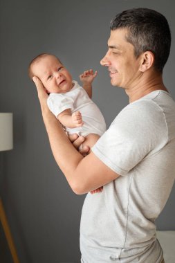 Beyaz tişörtlü, güler yüzlü beyaz bir adam yeni doğmuş bebeğini kollarında gri duvara yaslayıp küçük oğluyla mutlu vakit geçiriyor.
