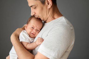 Beyaz tişörtlü genç bir adam, yeni doğmuş bebeğini kollarında tutarken beyaz tişörtlü, küçük oğlunu sevgiyle ve nazikçe öpüyor.