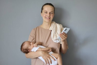 İyimser gülümseyen anne, kızı ya da oğlu bej tişört giymiş gri arka plan kadınına izole bir şekilde poz veriyor. Annesinin ev işlerini yaparken cep telefonu kullanıyor.