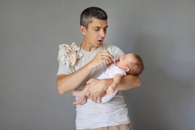Omuzlarında çocuk kıyafetleri olan sevecen bir baba. Gri arka planda izole edilmiş ağlayan bir bebeği tutuyor. Bebek emziğiyle oğlunu sakinleştirmeye çalışıyor.