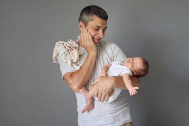 Sevecen, beyaz bir baba yeni doğmuş bebeğini kucağında tutuyor ve gizli çocuğuna hayranlık duyuyor. Gri arka planda yaşayan, çocuk elbiseli, ev işi yapan ve çocuğa bakan yakışıklı bir adam.