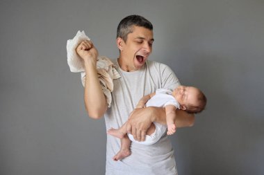 Aşırı mutlu, beyaz bir baba, yeni doğmuş bebeğini kucağında tutarak mutluluktan çığlık çığlığa bağırıyor. Gri arka planda izole edilmiş babalığının keyfini çıkarıyor.