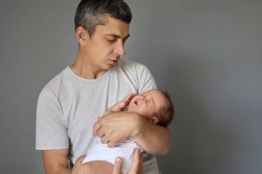 Ebeveynliğin ilk günleri. Sevimli, beyaz bir baba, yeni doğmuş bebeğini kollarında, aşk çocuğuyla, babasının ellerinde uyuyakalıyor. Gri arka planda tek başına.