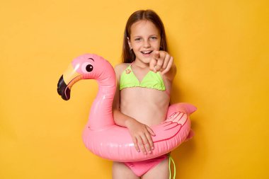 Pembe flamingo şişirilebilir yüzükte mayo giyen neşeli küçük kız kamerayı işaret ederek sarı arka planda izole edilmiş mutlu yüzünü seçiyor.
